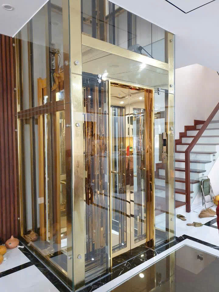 Thang máy kính khung inox mạ vàng lắp đặt trong nhà bên cạnh cầu thang bộ hiện đại